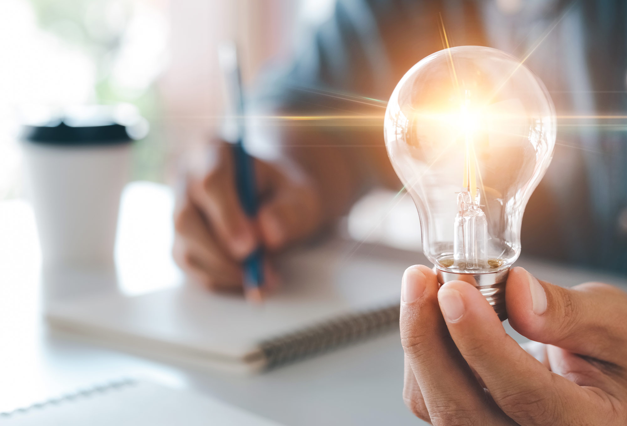 a left hand holds a glowing lightbulb in the focus in the foreground while the right hands writes in a notebook out of focus in the background